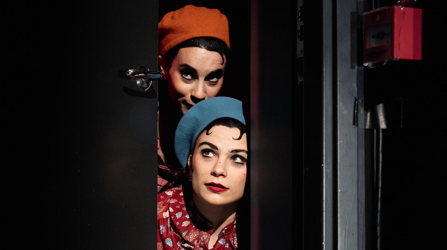 Två kvinnliga mimskådespelarstudenter tittar fram bakom en svart dörr. Från examensföreställningen Festfolk, 2022.