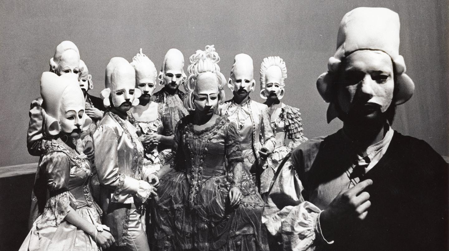 En grupp operasångare i halvmasker, med stora peruker i gips.