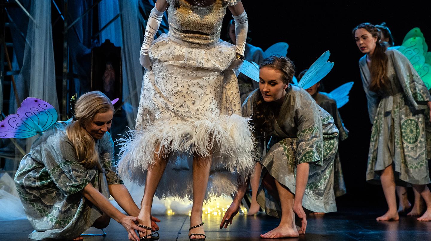 Fyra operastudenter på scen med änglavingar på ryggarna, varav en hjälper en annan att sätta på en sko. Från föreställningen En afton på Operan, 2022.