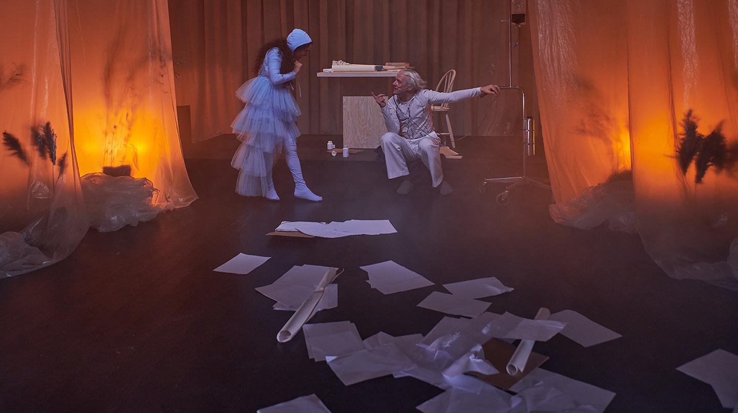 Två skådespelare i fonden och framför dem en massa papper utspridda på golvet. Från föreställningen Stormen - del av en scenkonstkurs 2022.