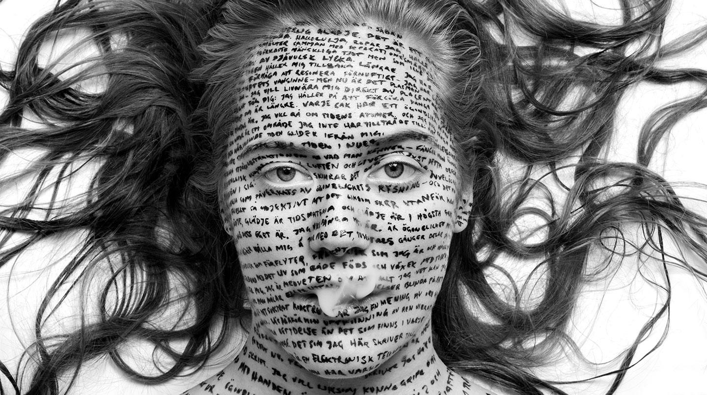 Ett kvinnoansikte med skriven text över hela ansiktet, håret utspritt och rök som kommer ur munnen. Från skådespelarstudenten Malou Zilliacus Departure-föreställning 2014.