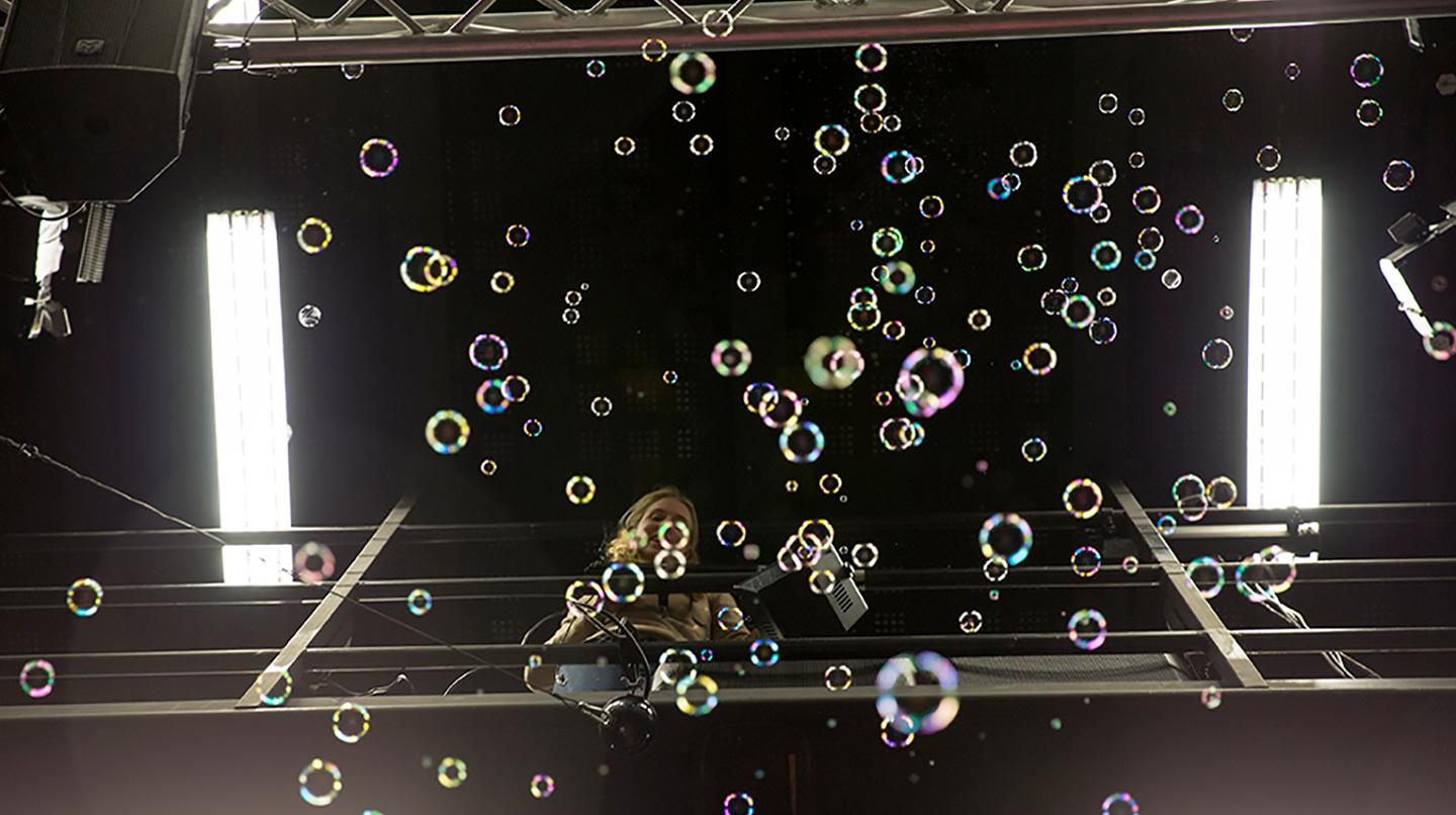 En scenkonststudent står högt upp på en ljusrigg och blåser såpbubblor.