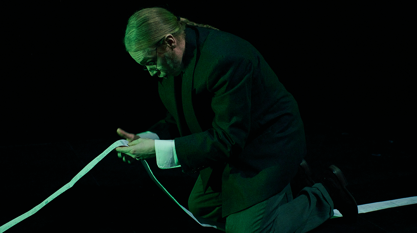 En man står på knä och läser på en meterlång pappersutskrift. Från forskarprojektet/operaföreställningen Don Pascuale, 2023.