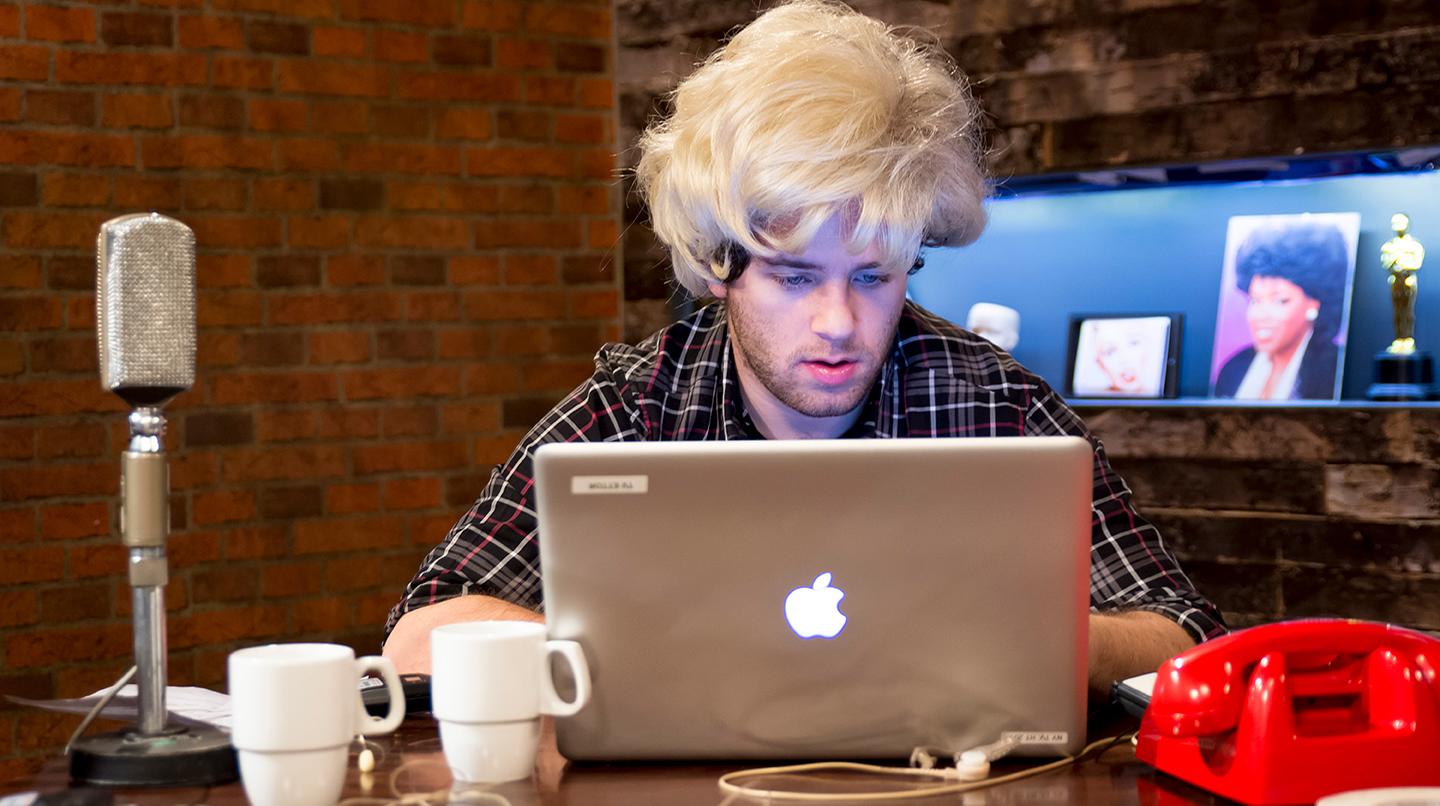 En tv-student i blond peruk, sitter vid ett skrivbord bakom en dator. Från kursen Tv-fabriken, 2015.