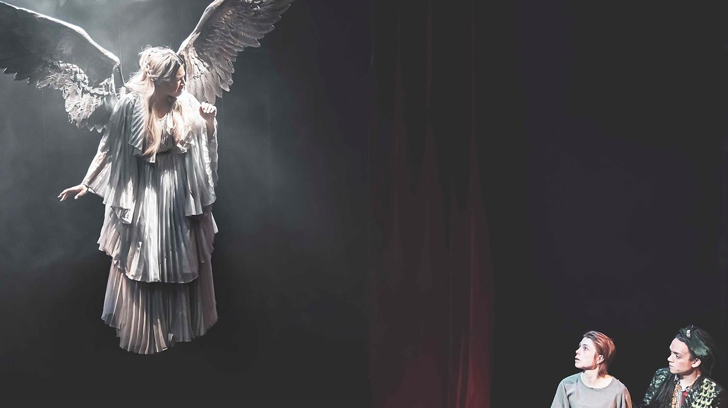 Två skådespelare sitter på golvet och tittar upp mot en ängel som hänger i taket. Från examensföreställningen Angels in America, skådespeleriklassen 2021.